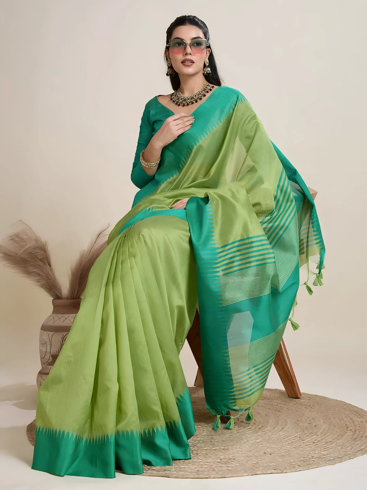 A woman models a striking two-toned Green Raw Silk Saree, transitioning between a light Pista Green and a deep Teal. The pallu contrasts dramatically in a wide band of Teal, featuring horizontal stripe and traditional weaving. She wears a matching short-sleeve teal blouse and accessorizes with a chunky dark gemstone necklace and large gold earrings, posing indoors.