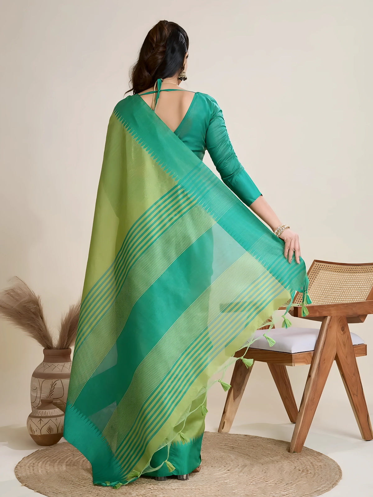 A woman models a striking two-toned Green Raw Silk Saree, transitioning between a light Pista Green and a deep Teal. The pallu contrasts dramatically in a wide band of Teal, featuring horizontal stripe and traditional weaving. She wears a matching short-sleeve teal blouse and accessorizes with a chunky dark gemstone necklace and large gold earrings, posing indoors.
