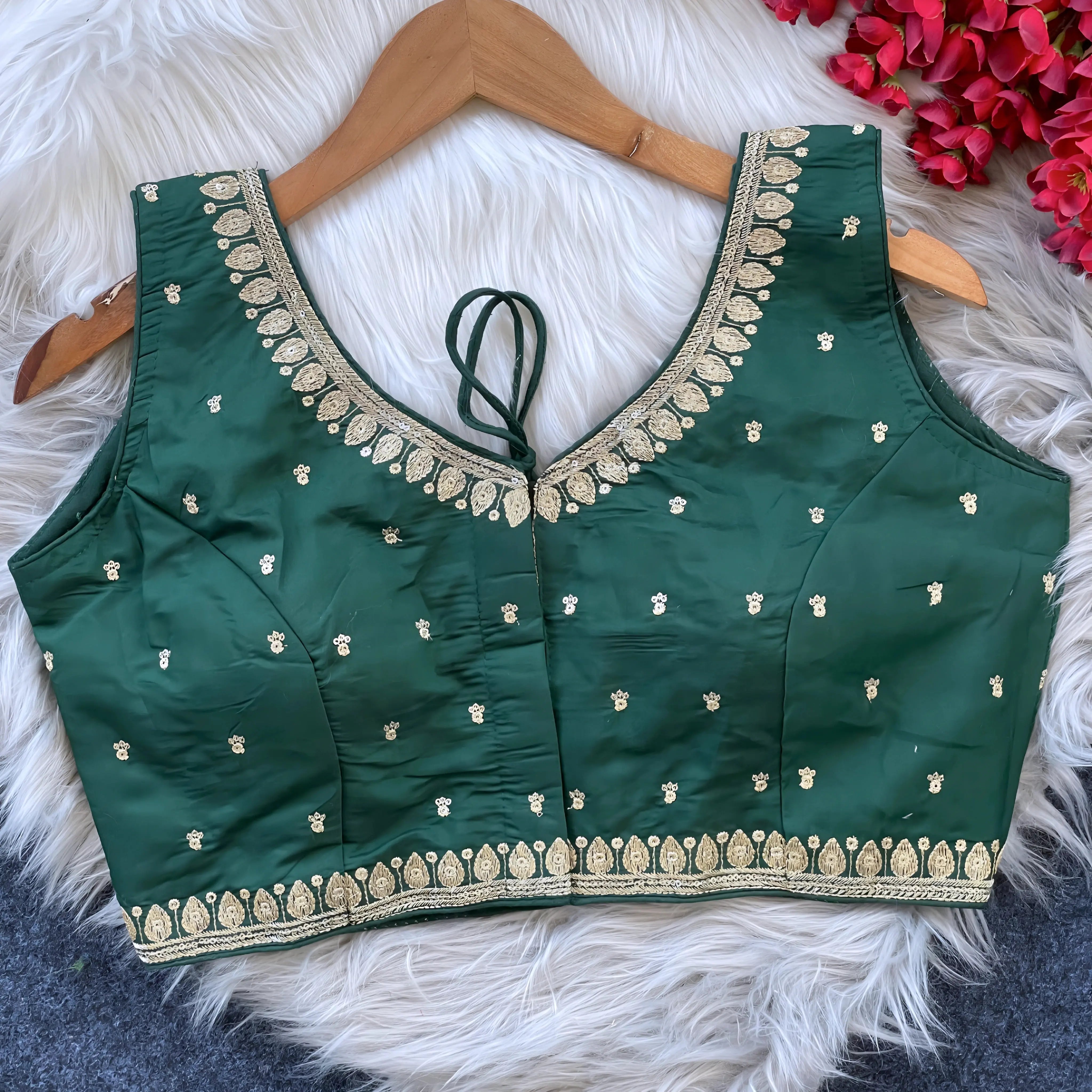 Green padded blouse with intricate gold embroidery along neckline and hem, featuring small embroidered motifs on the fabric, displayed on a wooden hanger over a white fur surface with red flowers nearby