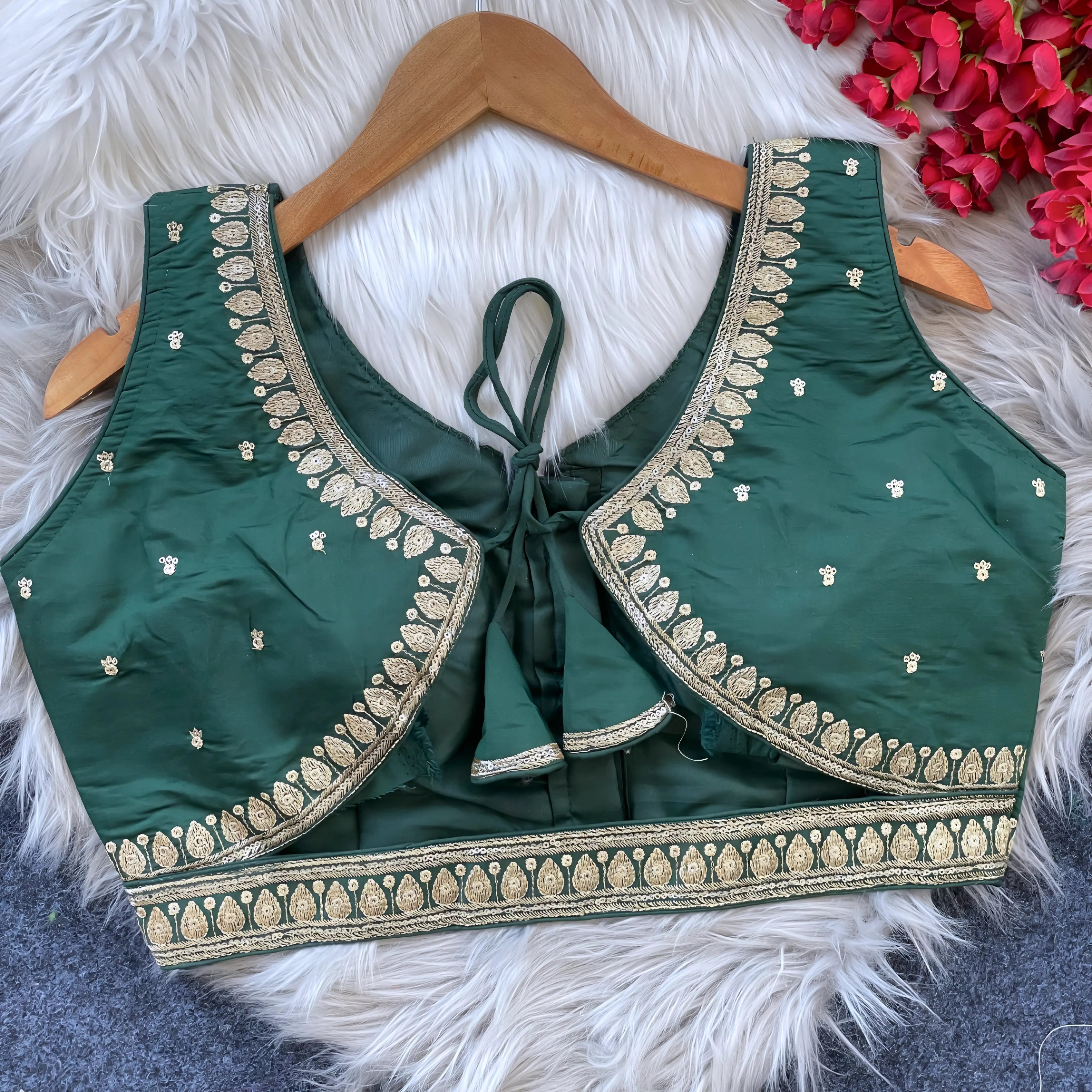 Green padded blouse with intricate gold embroidery along neckline and hem, featuring small embroidered motifs on the fabric, displayed on a wooden hanger over a white fur surface with red flowers nearby