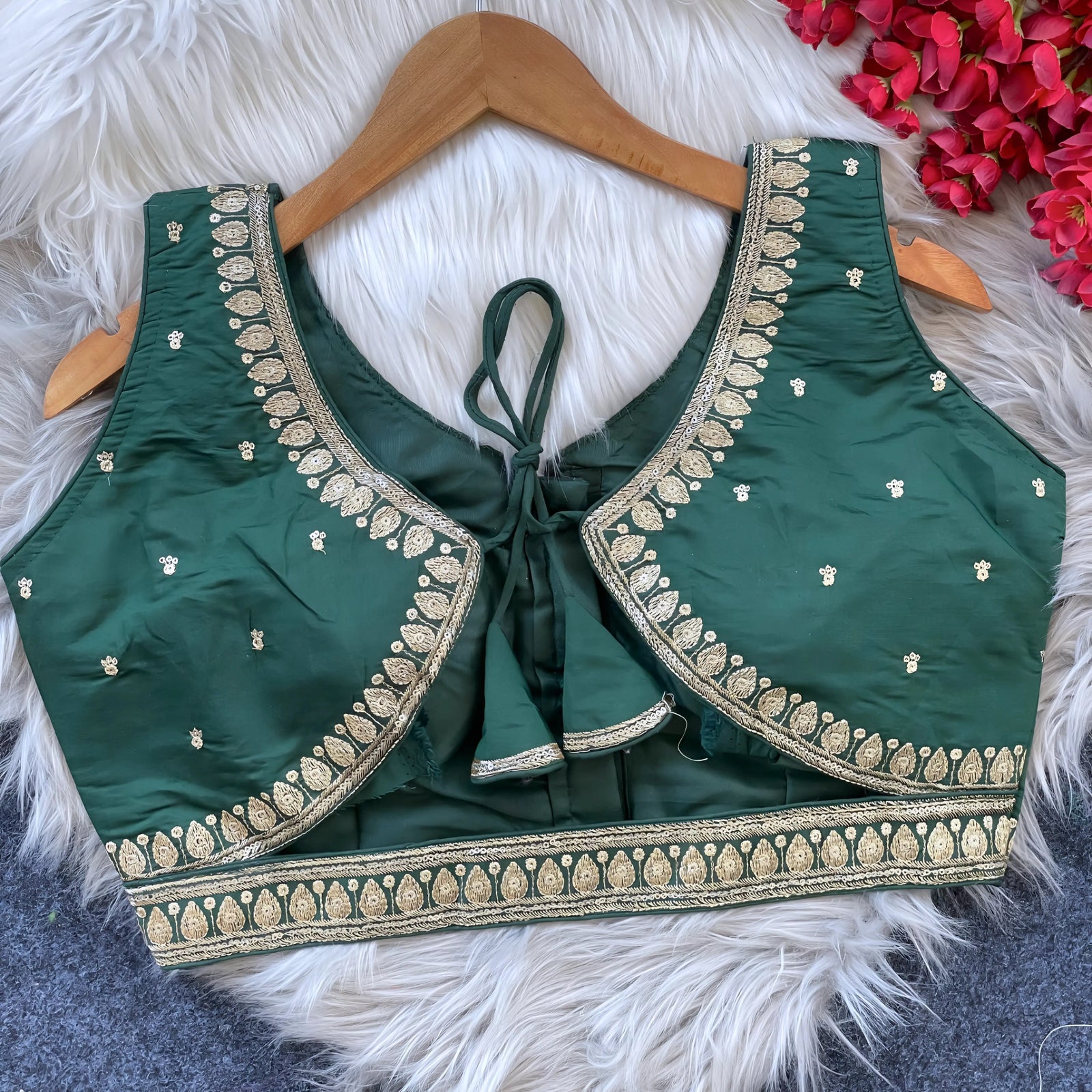 Green padded blouse with intricate gold embroidery along neckline and hem, featuring small embroidered motifs on the fabric, displayed on a wooden hanger over a white fur surface with red flowers nearby
