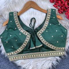 Green padded blouse with intricate gold embroidery along neckline and hem, featuring small embroidered motifs on the fabric, displayed on a wooden hanger over a white fur surface with red flowers nearby