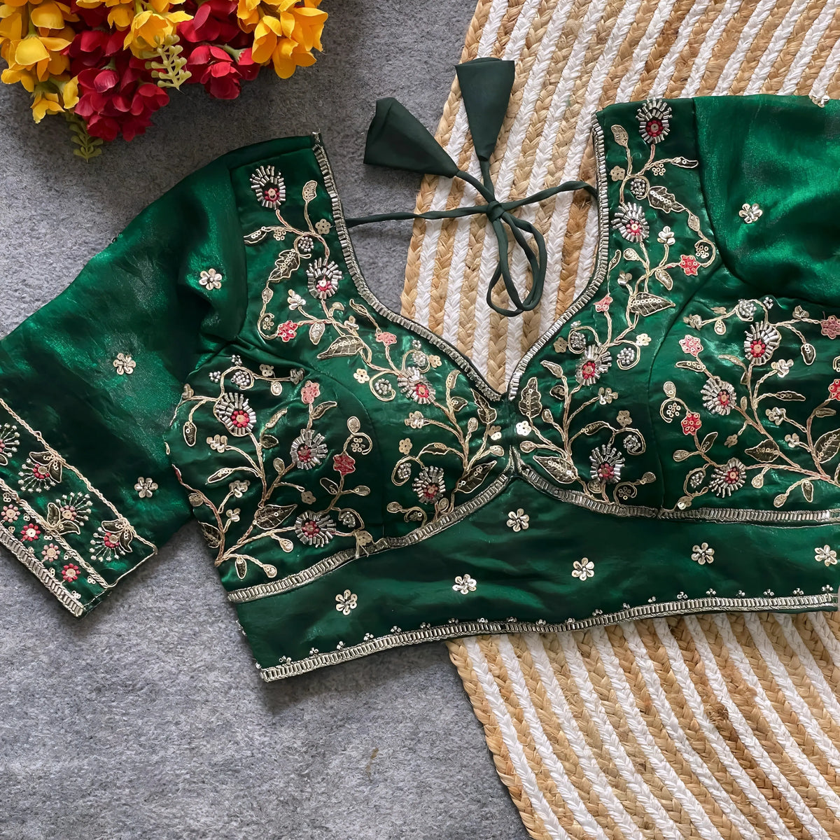 Green padded blouse with intricate multicolor floral embroidery and gold threadwork, featuring a tie-back detail, displayed on a grey surface with a woven mat and artificial yellow and red flowers in the background.