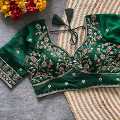Green padded blouse with intricate multicolor floral embroidery and gold threadwork, featuring a tie-back detail, displayed on a grey surface with a woven mat and artificial yellow and red flowers in the background.