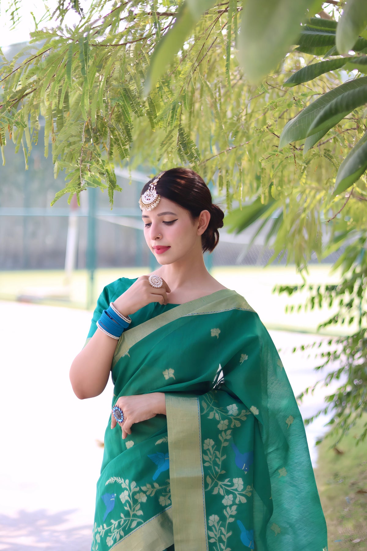 A woman models a vibrant Emerald Green Linen Silk Saree with delicate Zari woven floral and bird motifs. The saree has a wide, contrasting pale gold/pistachio green border. She wears bold blue bangles and a headpiece, posing outdoors.