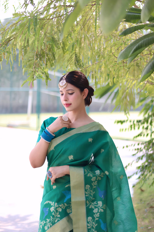 A woman models a vibrant Emerald Green Linen Silk Saree with delicate Zari woven floral and bird motifs. The saree has a wide, contrasting pale gold/pistachio green border. She wears bold blue bangles and a headpiece, posing outdoors.
