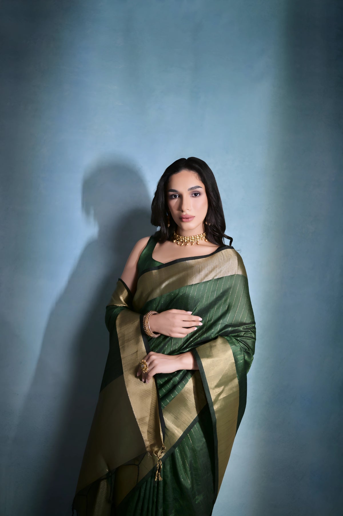 A woman models a striking Dark Green Banarasi Raw Silk Saree. The saree body features subtle gold Zari stripes and is framed by an extremely wide, luxurious metallic gold Zari border. She wears a sleeveless dark blouse and traditional gold Kundan jewelry, posing indoors.