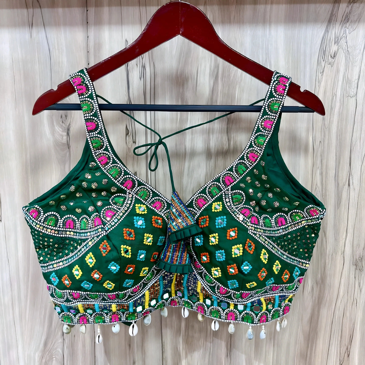  Traditional green Gujarati-style blouse featuring vibrant multicolor geometric embroidery in yellow, pink, blue, and orange, accented with intricate mirror work and white cowrie shell trim along the hem. The design includes wide embroidered straps, structured cups, decorative front detail, and tasseled tie-back, displayed on a wooden hanger against a light wood panel background.