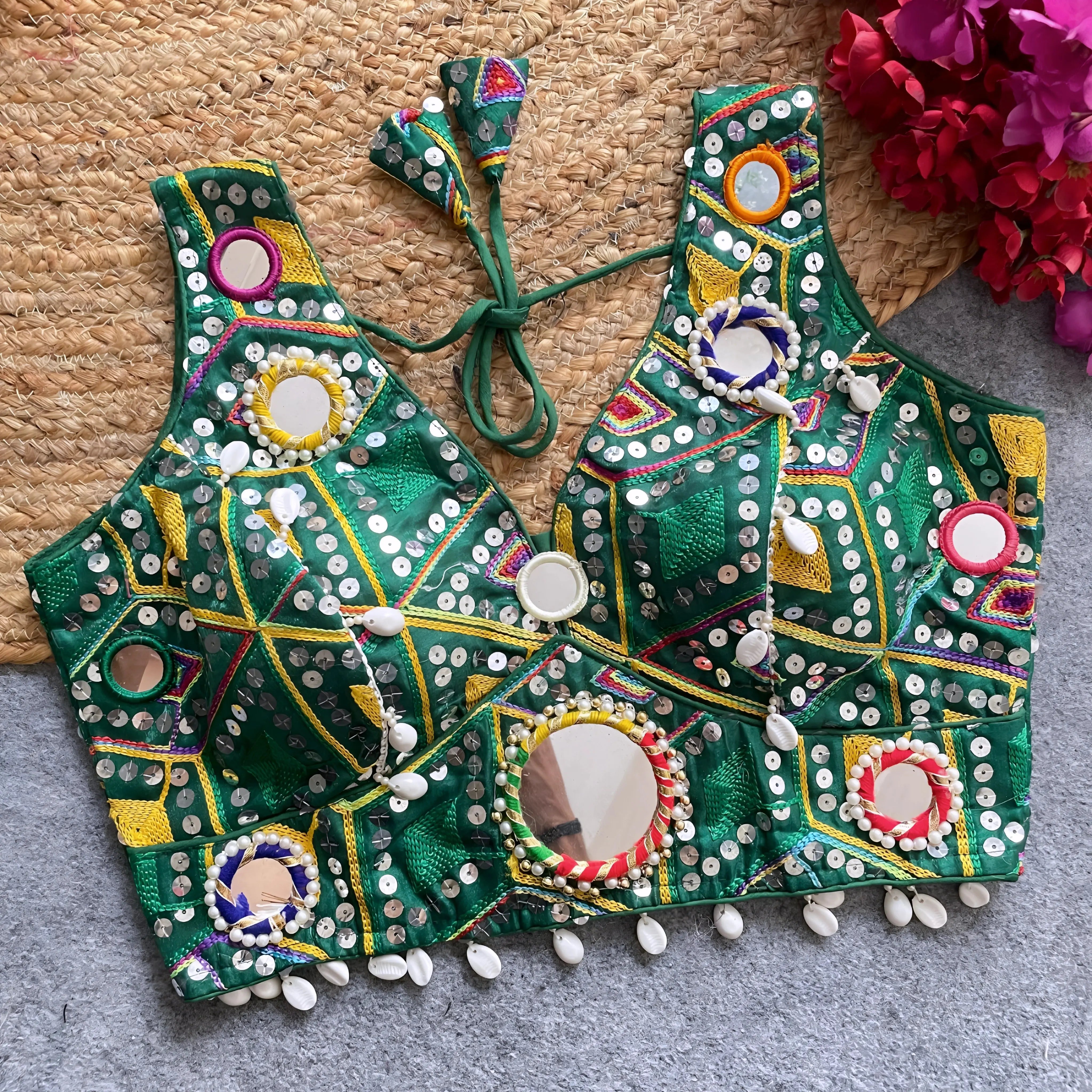 Traditional green Gujarati-style blouse adorned with intricate multicolor geometric embroidery, large circular mirrors bordered with colorful beads, and decorative white cowrie shell trim along the hem. The blouse features wide straps, detailed mirror work, and tasseled tie-back detail, displayed flat on a grey carpet with a woven mat and vibrant fuchsia artificial flowers.