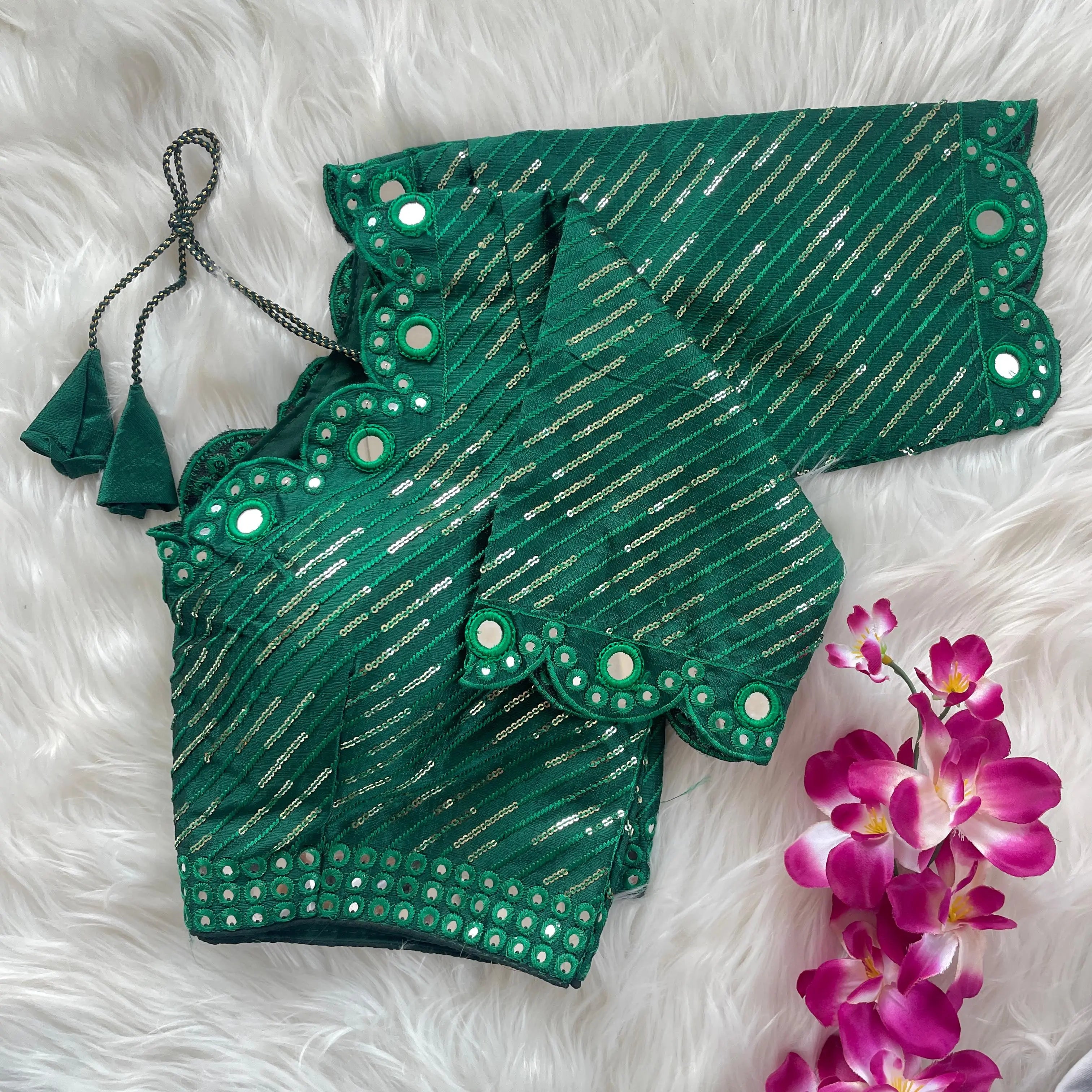 Green blouse with silver sequin stripes, scalloped neckline, and mirror work detailing, displayed on a textured grey and white fur background with a small potted plant nearby.