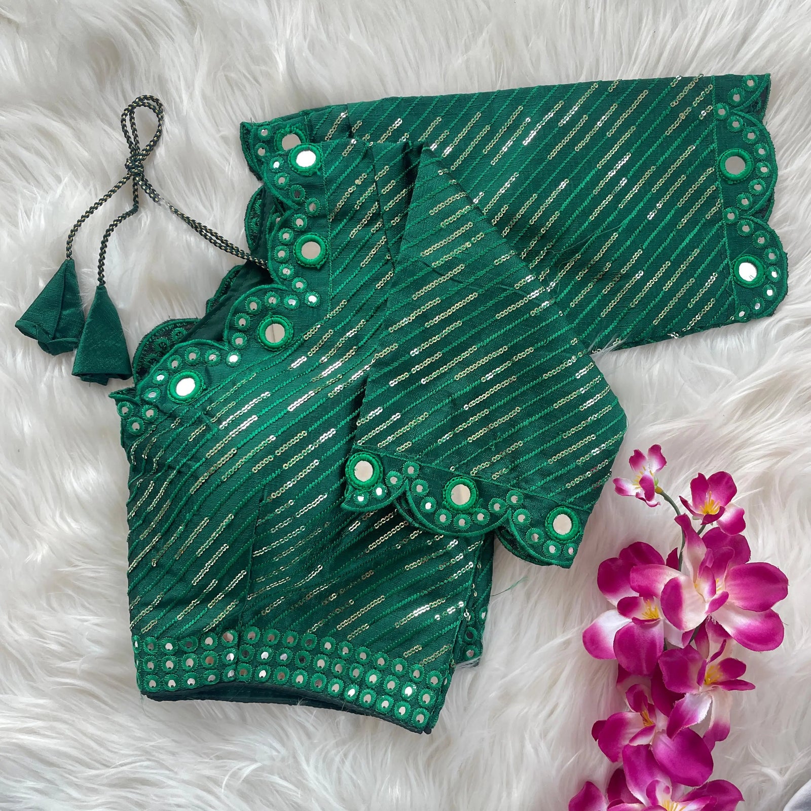 Green blouse with silver sequin stripes, scalloped neckline, and mirror work detailing, displayed on a textured grey and white fur background with a small potted plant nearby.