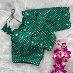 Green blouse with silver sequin stripes, scalloped neckline, and mirror work detailing, displayed on a textured grey and white fur background with a small potted plant nearby.