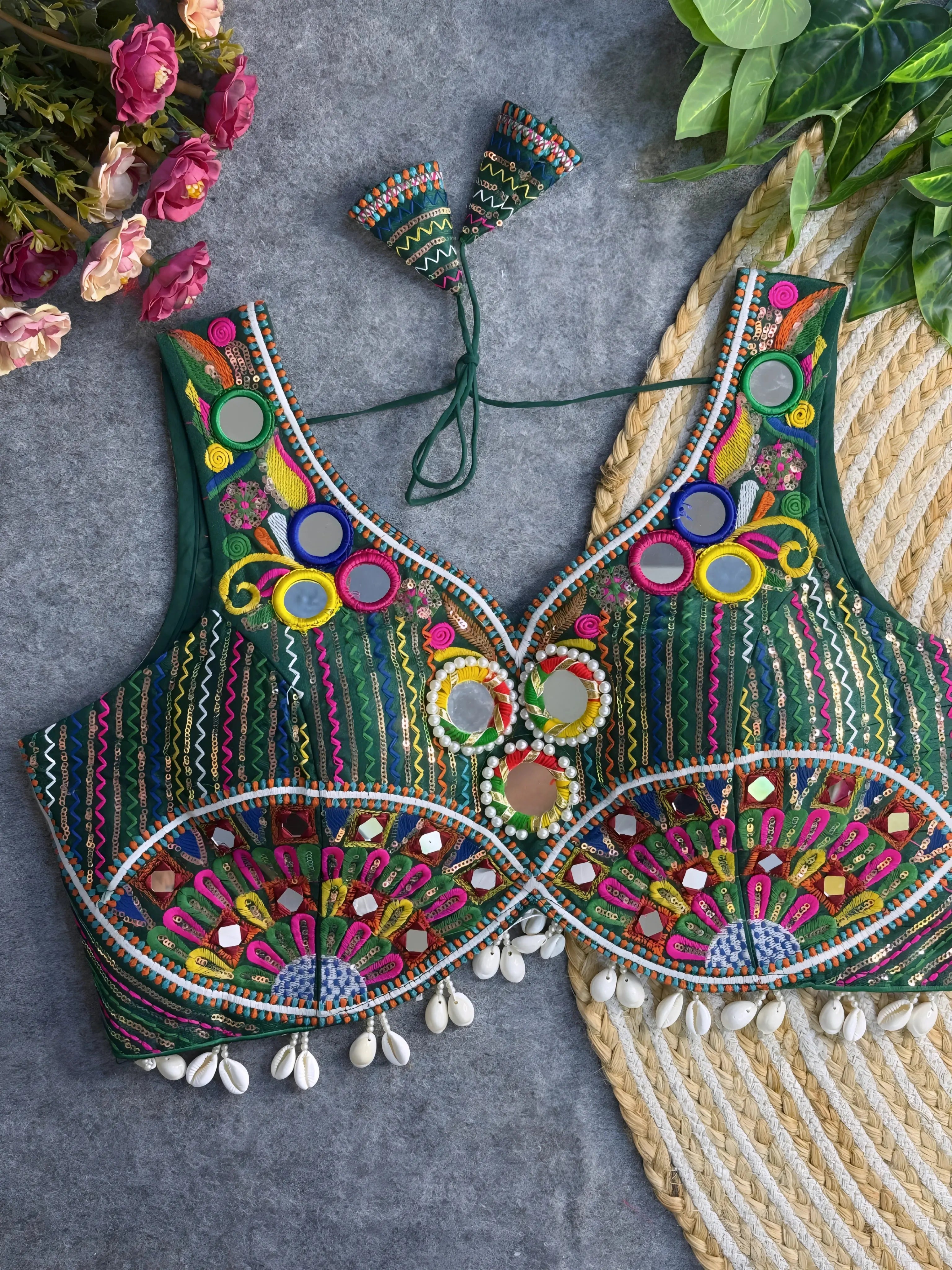 Traditional green embroidered blouse featuring vibrant multicolor mirror work, intricate thread embroidery, and shell tassel detailing along the hem, displayed on a flat surface with floral and leafy decor.