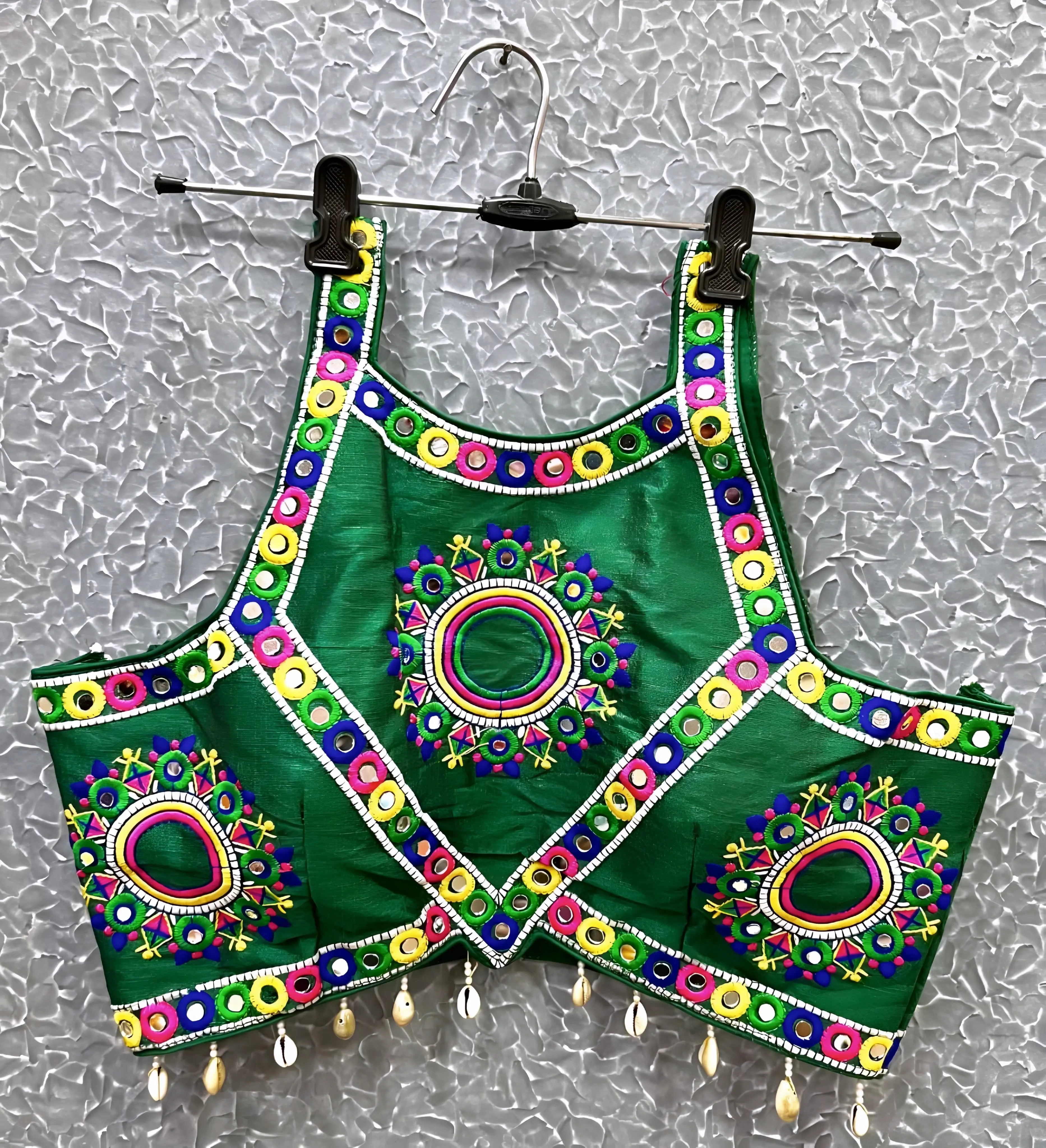 Green blouse featuring vibrant circular and oval mirror work with traditional Kutch multicolor embroidery, accented by geometric borders and shell tassel drops along the hem. Displayed on a hanger against a textured silver background, perfectly showcasing its festive and culturally inspired ethnic design.
