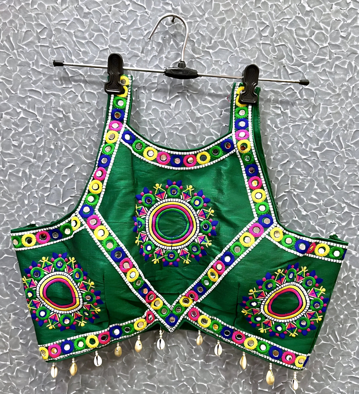 Green blouse featuring vibrant circular and oval mirror work with traditional Kutch multicolor embroidery, accented by geometric borders and shell tassel drops along the hem. Displayed on a hanger against a textured silver background, perfectly showcasing its festive and culturally inspired ethnic design.