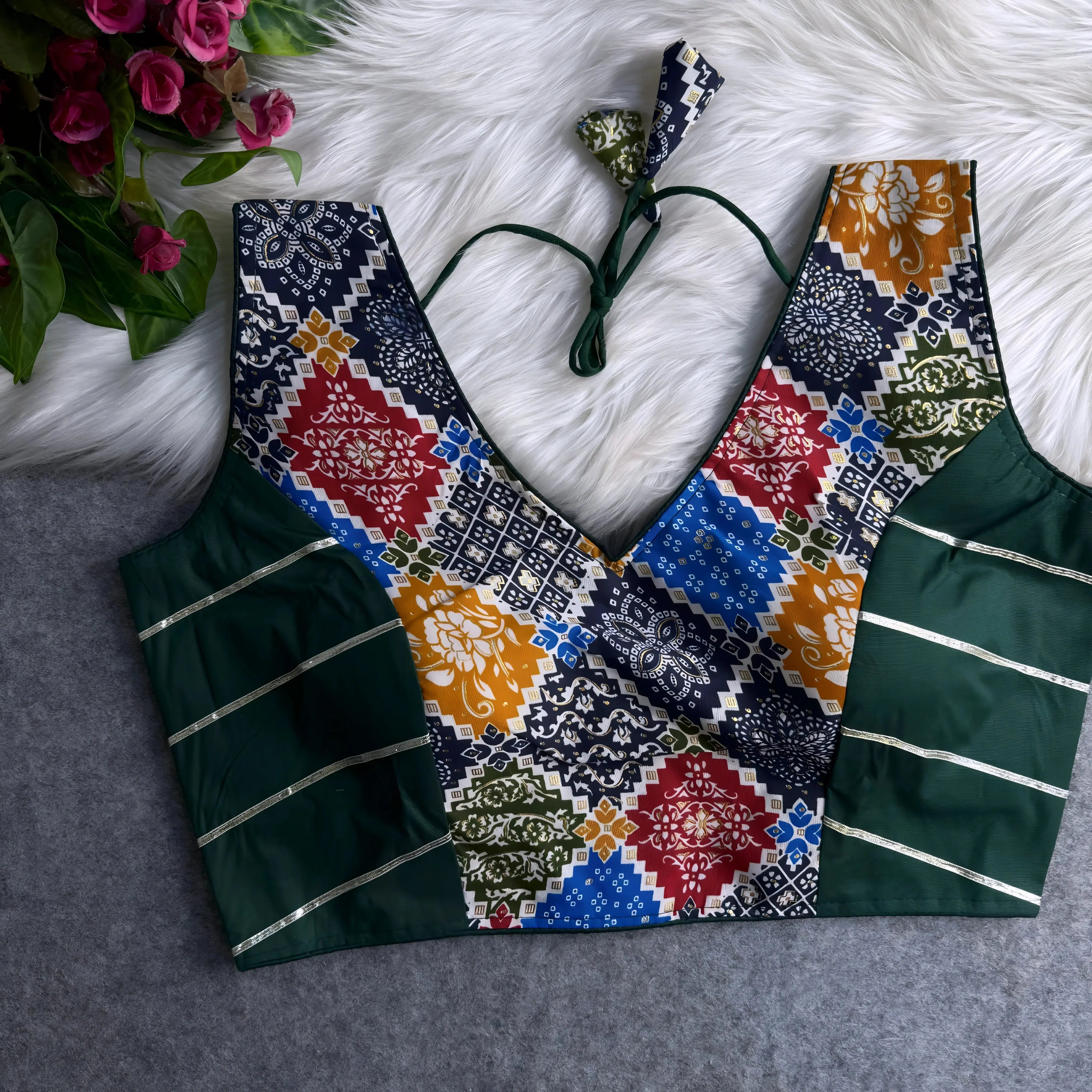 Traditional blouse featuring green side panels with silver stripes and a multicolor Ajrak-inspired block print on the front, showcasing geometric and floral motifs in blue, mustard, red, yellow, and green. The design includes a deep V-neckline, tasseled tie-back detail, and is displayed flat on a white fur background with artificial pink flowers in the corner.