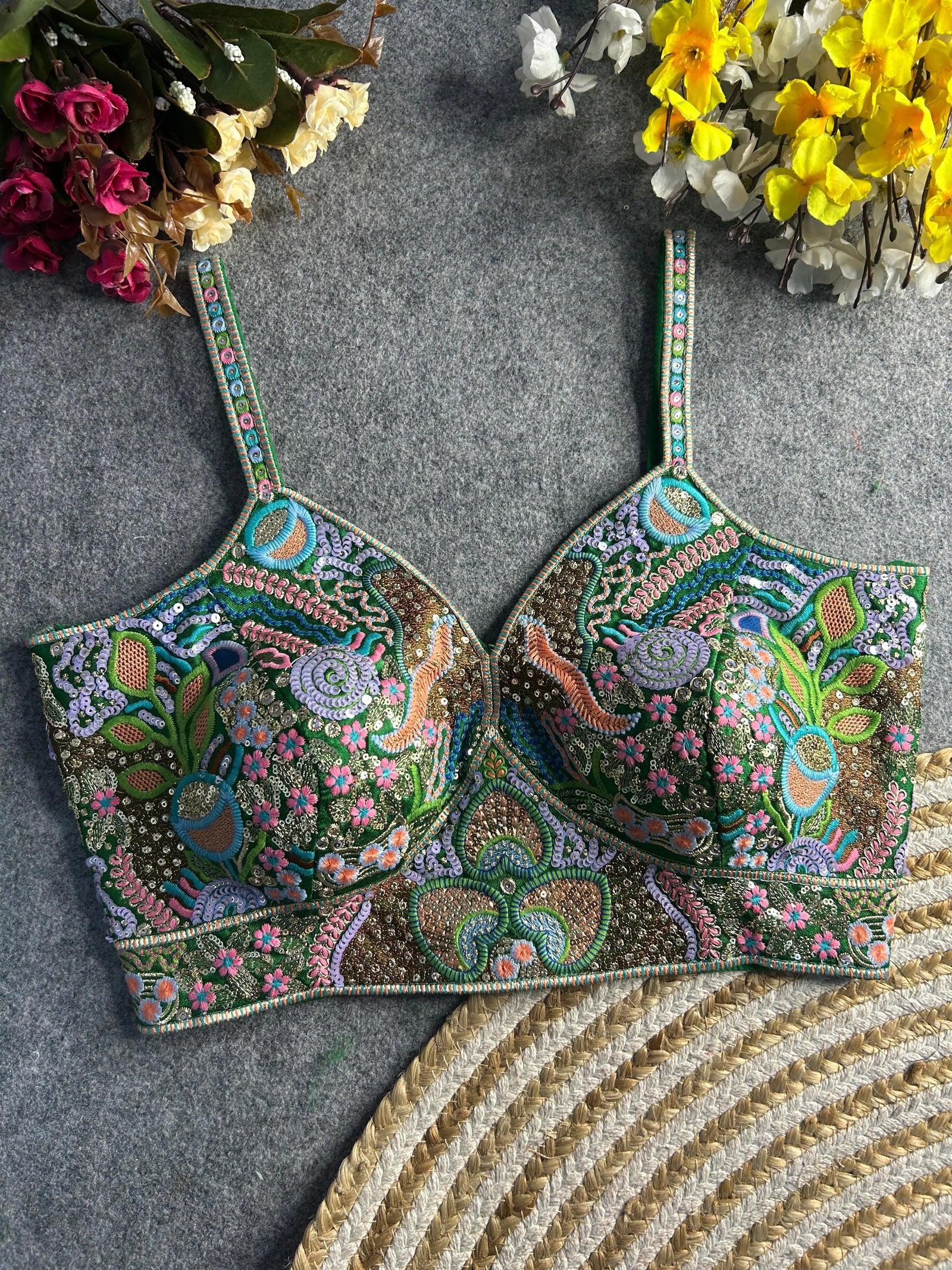 Traditional green bustier-style blouse featuring vibrant multicolor Madhubani-inspired embroidery with intricate floral, paisley, peacock, and geometric motifs, embellished with sequins and beads, structured cups, and adjustable straps, displayed flat on a textured grey surface with artificial pink, yellow, and white flowers in the corners.