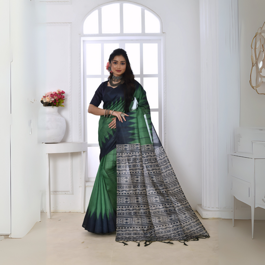 A woman models a dramatic Two-Toned Green Soft Tussar Silk Saree. The saree features bold black geometric patterns along the border and a pallu with dense abstract black, grey, and white tribal-style print. She wears a black blouse and chunky oxidized silver jewelry, posing indoors by a window.