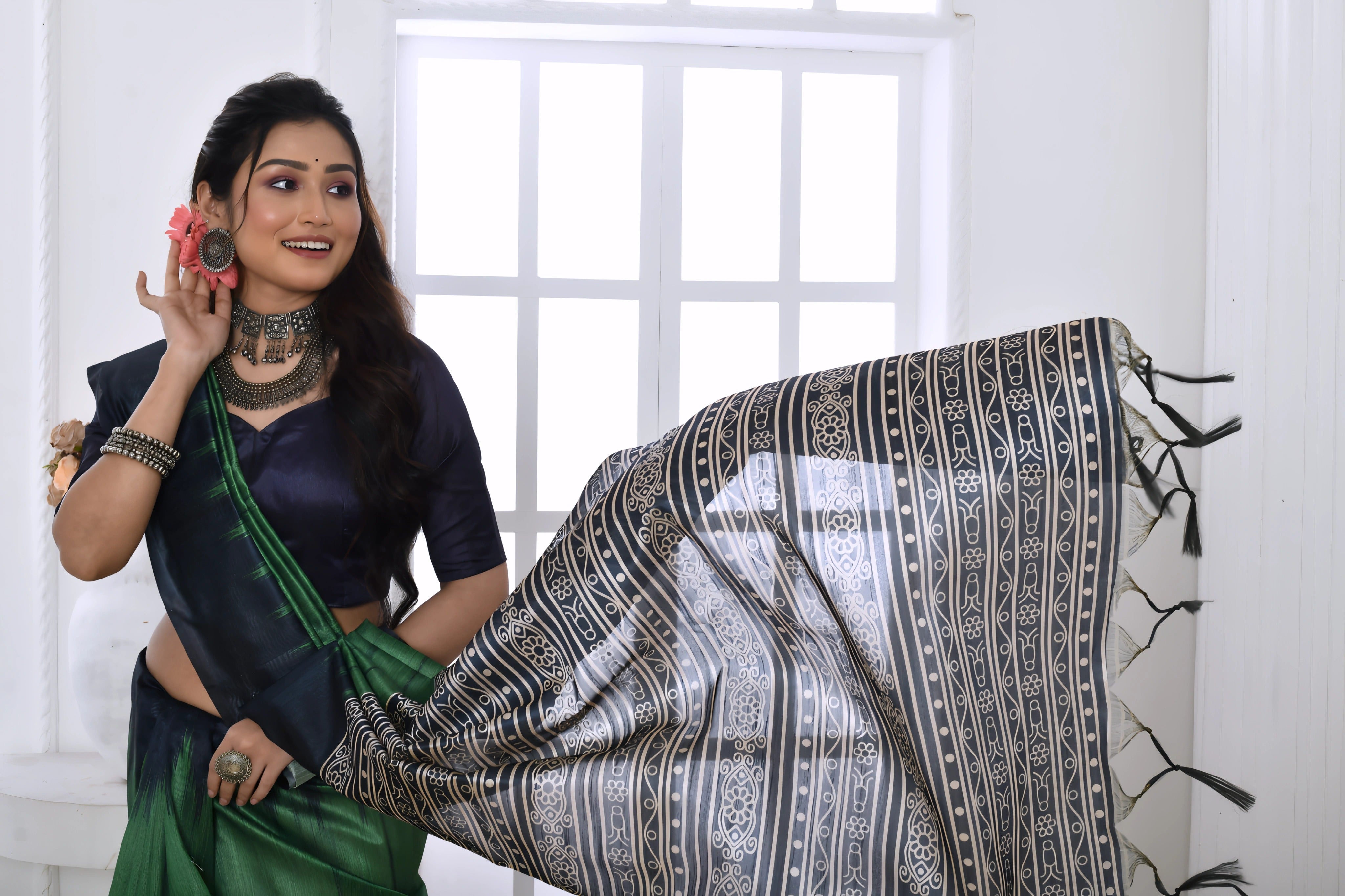 A woman models a dramatic Two-Toned Green Soft Tussar Silk Saree. The saree features bold black geometric patterns along the border and a pallu with dense abstract black, grey, and white tribal-style print. She wears a black blouse and chunky oxidized silver jewelry, posing indoors by a window.