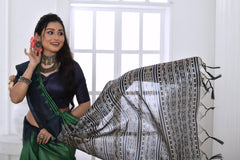 A woman models a dramatic Two-Toned Green Soft Tussar Silk Saree. The saree features bold black geometric patterns along the border and a pallu with dense abstract black, grey, and white tribal-style print. She wears a black blouse and chunky oxidized silver jewelry, posing indoors by a window.