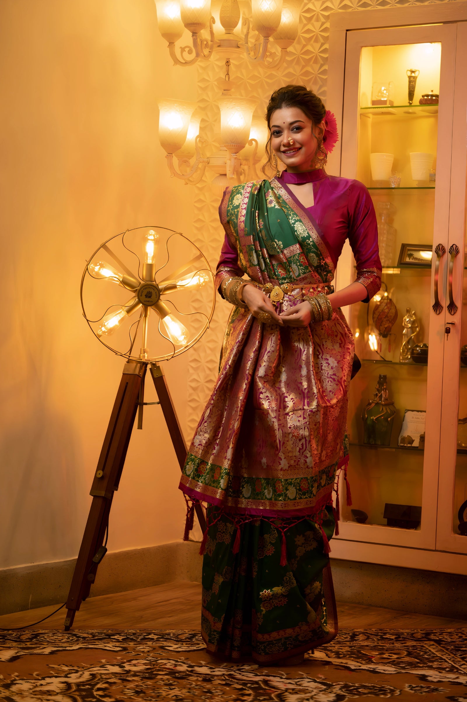 A woman models a luxurious Emerald Green Soft Banarasi Silk Saree. The saree features dense gold Zari weaving and a striking Fuchsia Pink contrast border/edging. She wears a high-neck, long-sleeve magenta blouse and gold jewelry, sitting in an opulent, dramatically lit setting.
