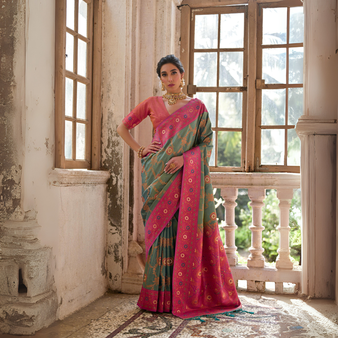 A woman models a striking Dusty Light Green Banarasi Silk Saree with subtle Zari weaving. The saree has a wide, ornate Fuchsia Pink border and pallu featuring dense traditional geometric Zari weaving. She wears a coral pink blouse and heavy gold Kundan jewelry.