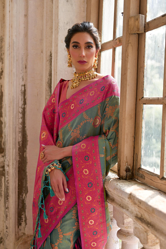 A woman models a striking Dusty Light Green Banarasi Silk Saree with subtle Zari weaving. The saree has a wide, ornate Fuchsia Pink border and pallu featuring dense traditional geometric Zari weaving. She wears a coral pink blouse and heavy gold Kundan jewelry.