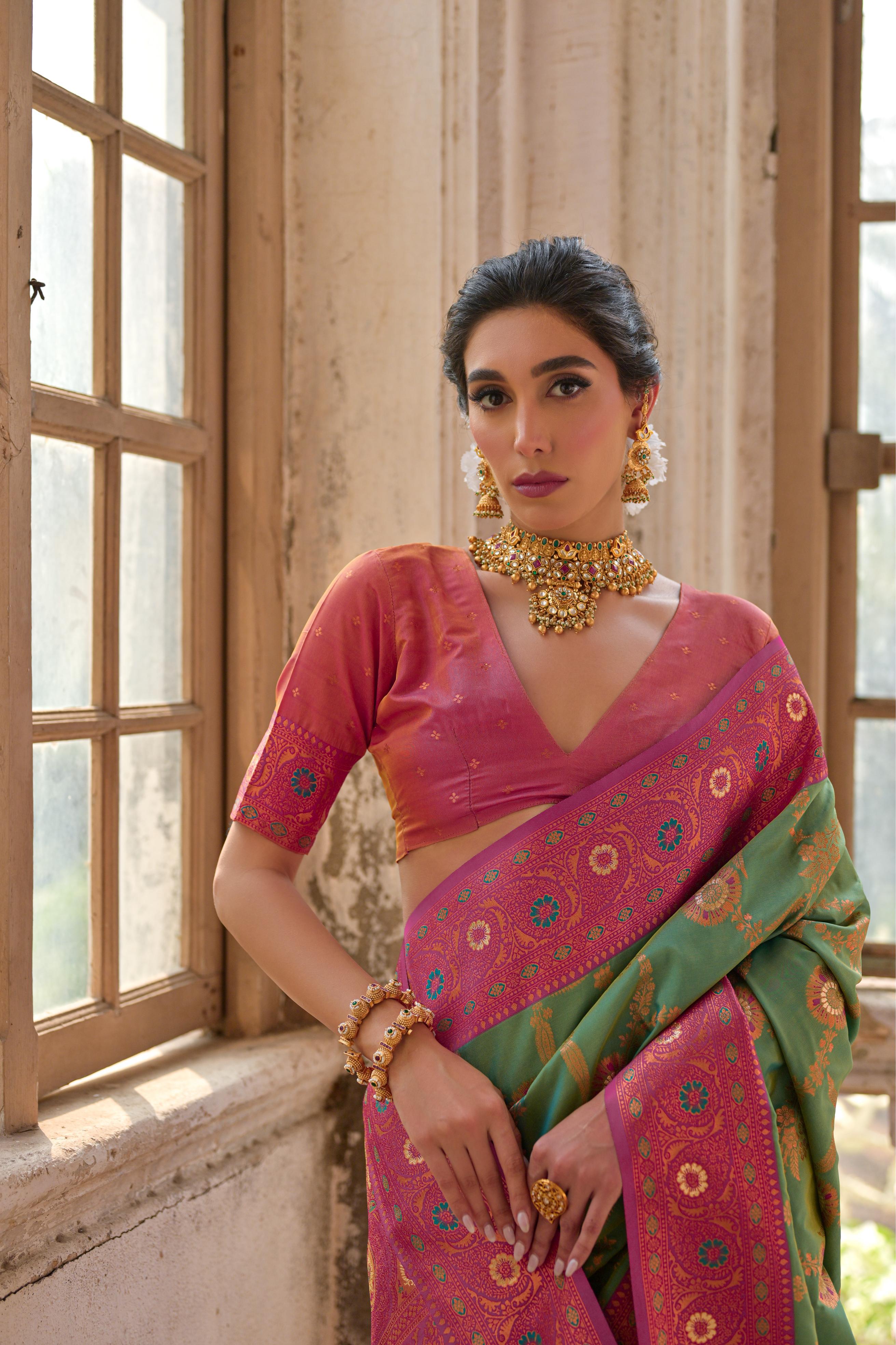A woman models a striking Moss Green Banarasi Silk Saree with gold Zari weaving. The saree has a wide, ornate Fuchsia Pink border and pallu featuring traditional geometric Zari weaving. She wears a pink and gold blouse and heavy gold Kundan jewelry.