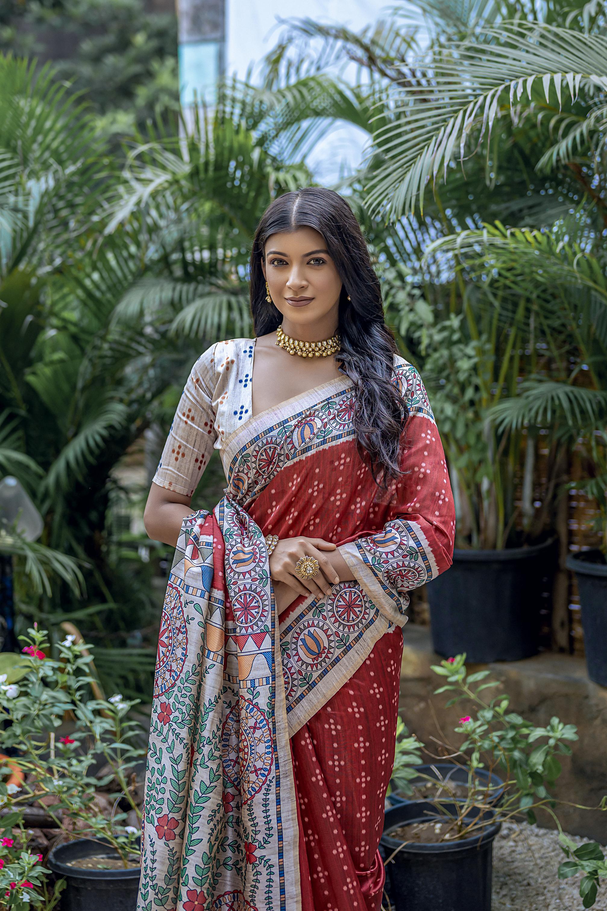 A woman models a vibrant Terracotta Red Tussar Silk Saree with small scattered prints. The pallu features a wide, elaborate panel with intricate Madhubani-style folk art prints in white, red, blue, and green. She wears a cream/gold embellished blouse and a gold Kundan necklace.