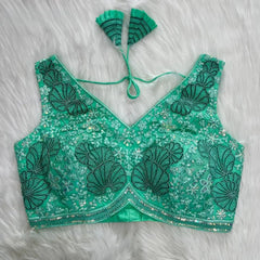  Aqua green blouse featuring intricate shell-shaped embroidery, sparkling sequin and bead embellishments, floral motifs, and a geometric border, with a deep V-neck, structured cups, and pleated fan-shaped tasseled tie-back detail, displayed flat on a white fur background.