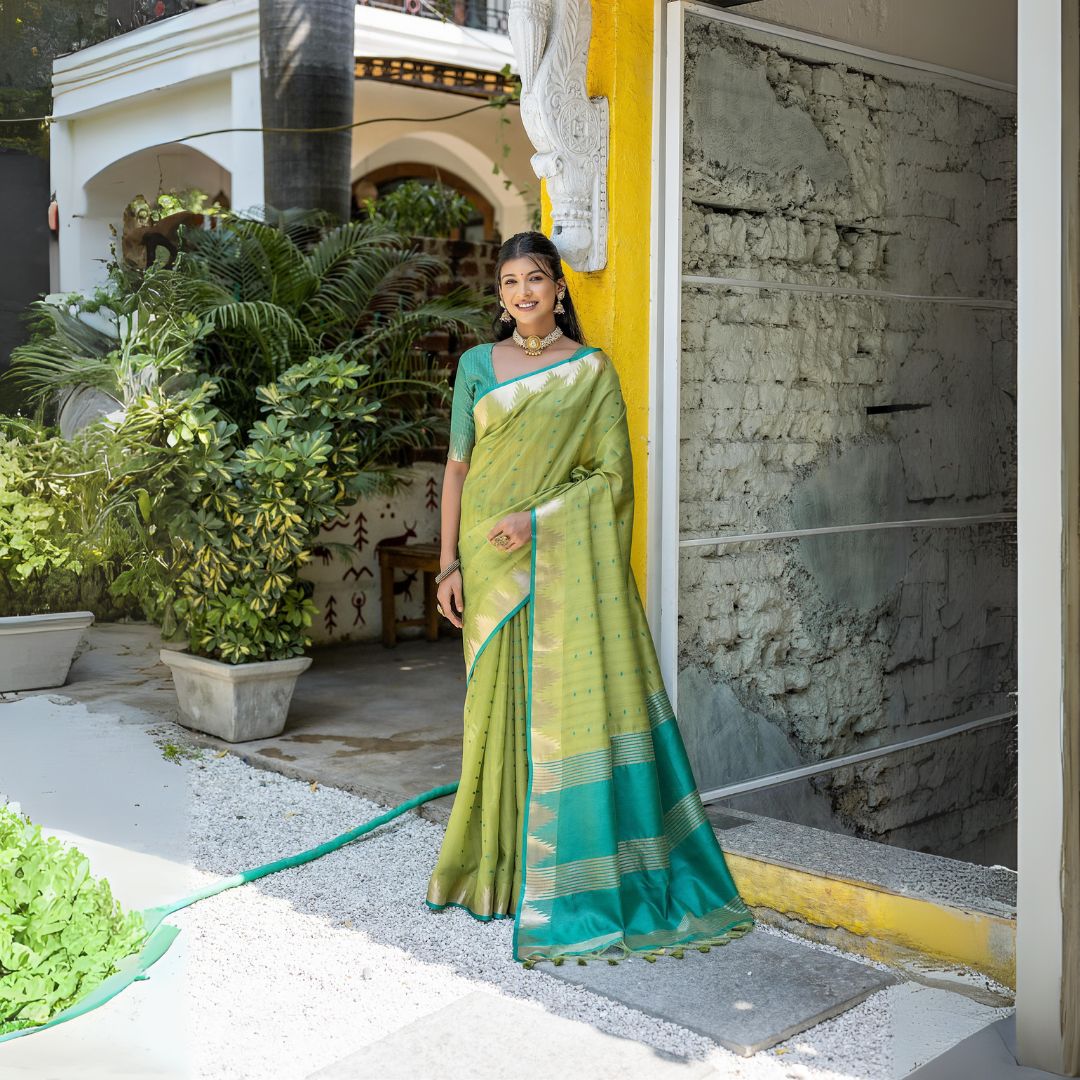 A woman models a vibrant Pista Green Tussar Silk Saree with small woven motifs. The saree has a contrasting peacock blue/teal pallu featuring wide gold Zari stripes. She wears a teal blouse and gold Kundan jewelry, posing in an outdoor courtyard.