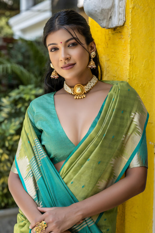 A woman models a vibrant Pista Green Tussar Silk Saree with small woven motifs. The saree has a contrasting peacock blue/teal pallu featuring wide gold Zari stripes. She wears a teal blouse and gold Kundan jewelry, posing in an outdoor courtyard.