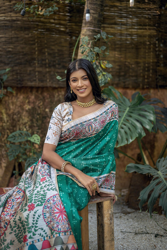 A woman models a vibrant Sea Green Soft Tussar Silk Saree. The pallu features a wide, elaborate panel with intricate Madhubani-style folk art prints in white, pink, and red, framed by a thin gold border. She wears a cream/gold blouse and a gold choker necklace.