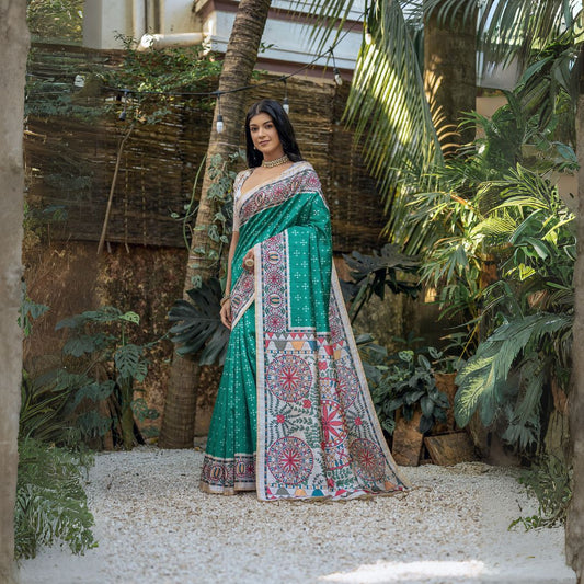 A woman models a vibrant Sea Green Soft Tussar Silk Saree. The pallu features a wide, elaborate panel with intricate Madhubani-style folk art prints in white, pink, and red, framed by a thin gold border. She wears a cream/gold blouse and a gold choker necklace.