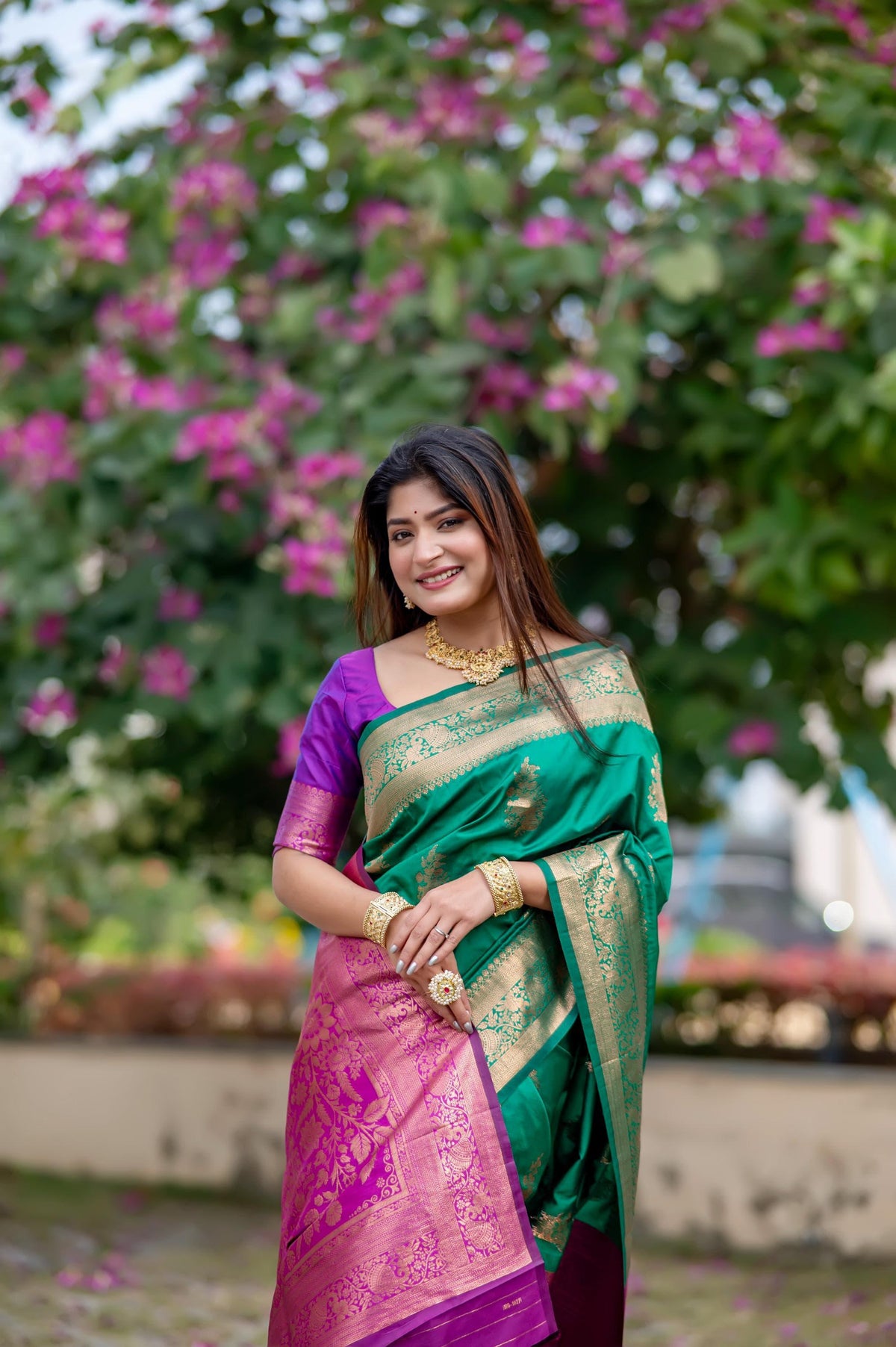 Woman in green zari work saree with purple blouse and gold jewelry posing in front of blooming pink flowers