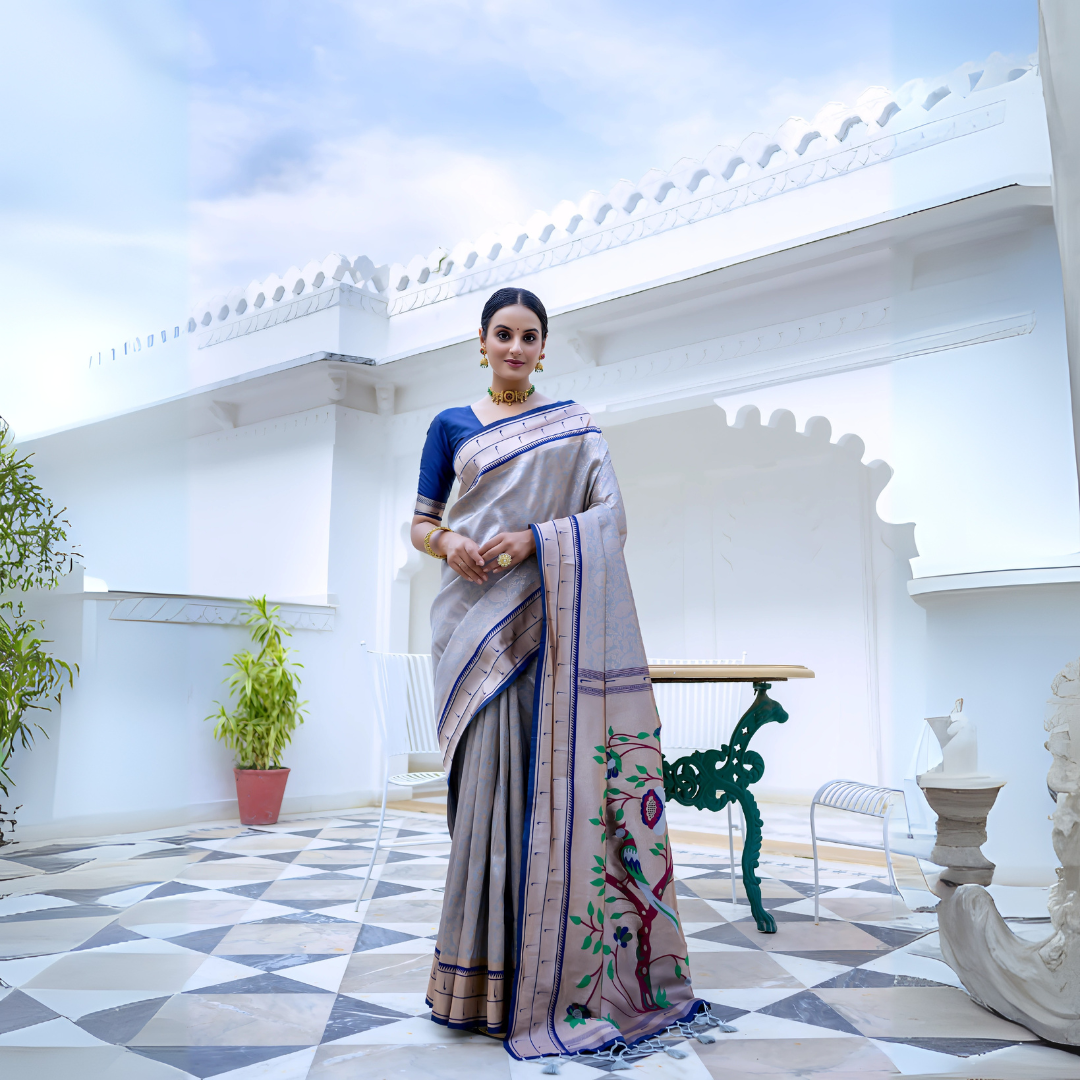 A woman models an elegant Light Gray Soft Silk Saree. The saree features a striking deep navy blue contrast border accented with gold Zari. The pallu is a focal point, featuring rich, artistic Paithani-style print or weaving of colorful bird-on-branch motifs. She wears a navy blue blouse and traditional gold jewelry with green accents, posing on an ornate white terrace.