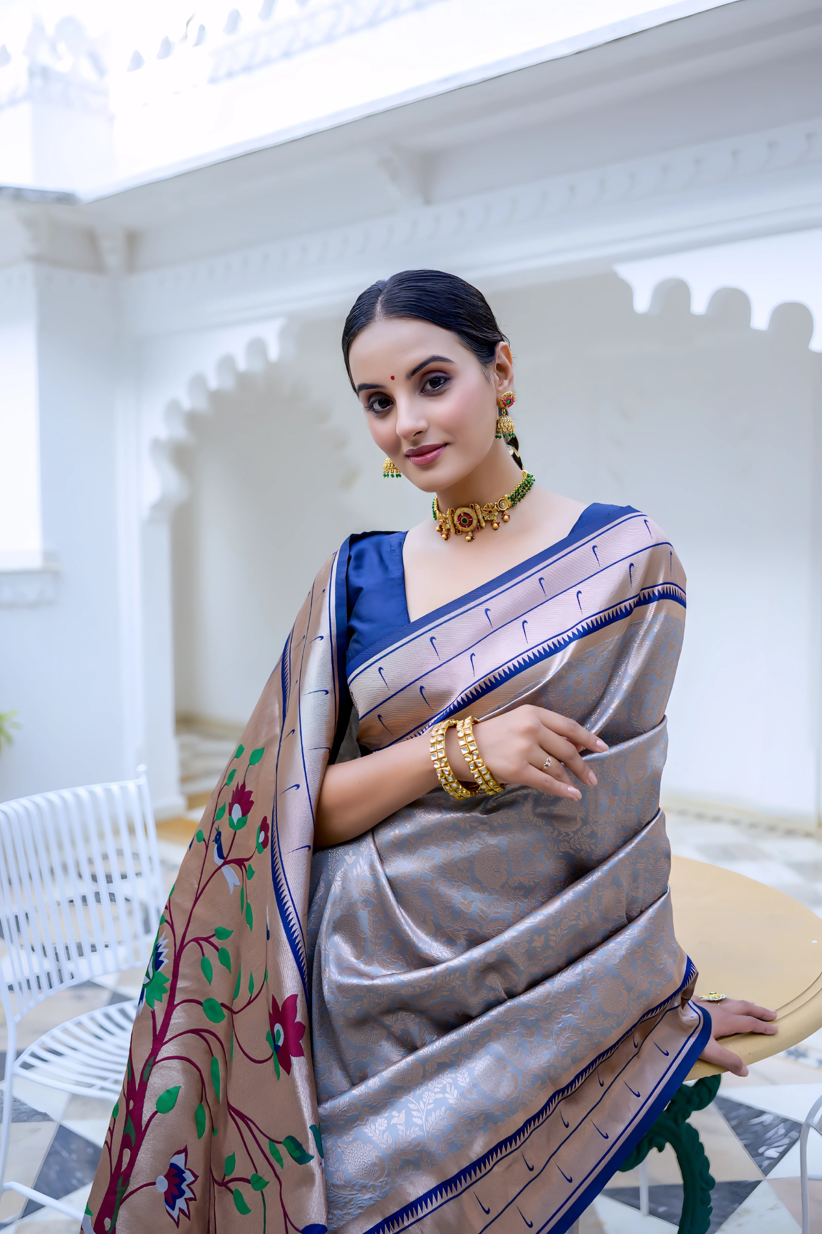 A woman models an elegant Light Gray Soft Silk Saree. The saree features a striking deep navy blue contrast border accented with gold Zari. The pallu is a focal point, featuring rich, artistic Paithani-style print or weaving of colorful bird-on-branch motifs. She wears a navy blue blouse and traditional gold jewelry with green accents, posing on an ornate white terrace.