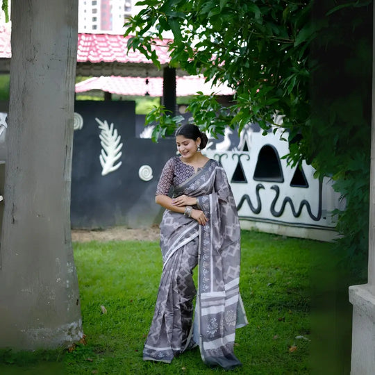 A woman models a striking Charcoal Grey Soft Cotton Saree featuring an all-over digital block print of geometric and abstract motifs in white. She wears a printed grey blouse and stacked silver bangles, posing outdoors against a stylized black and white wall.