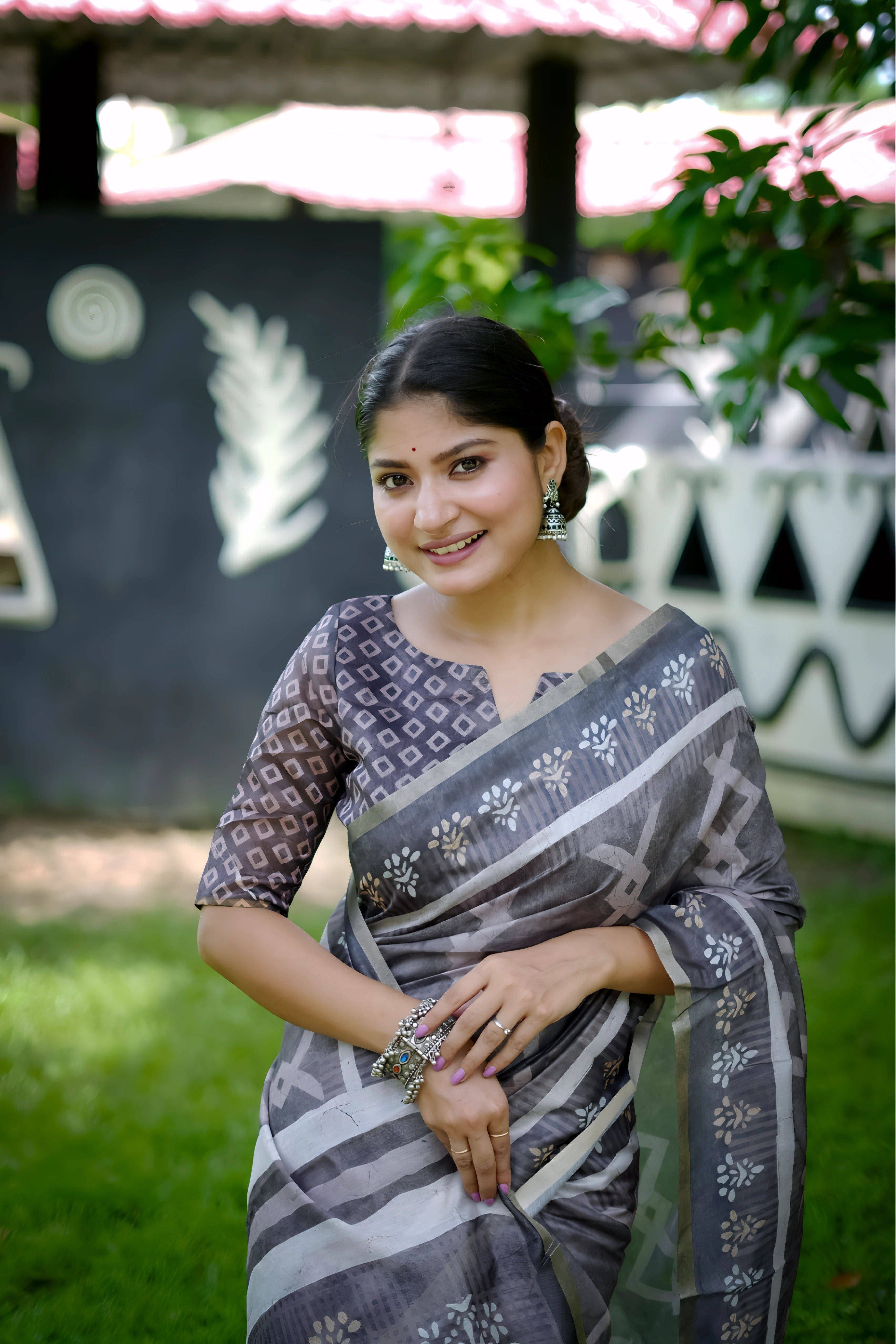 A woman models a striking Charcoal Grey Soft Cotton Saree featuring an all-over digital block print of geometric and abstract motifs in white. She wears a printed grey blouse and stacked silver bangles, posing outdoors against a stylized black and white wall.