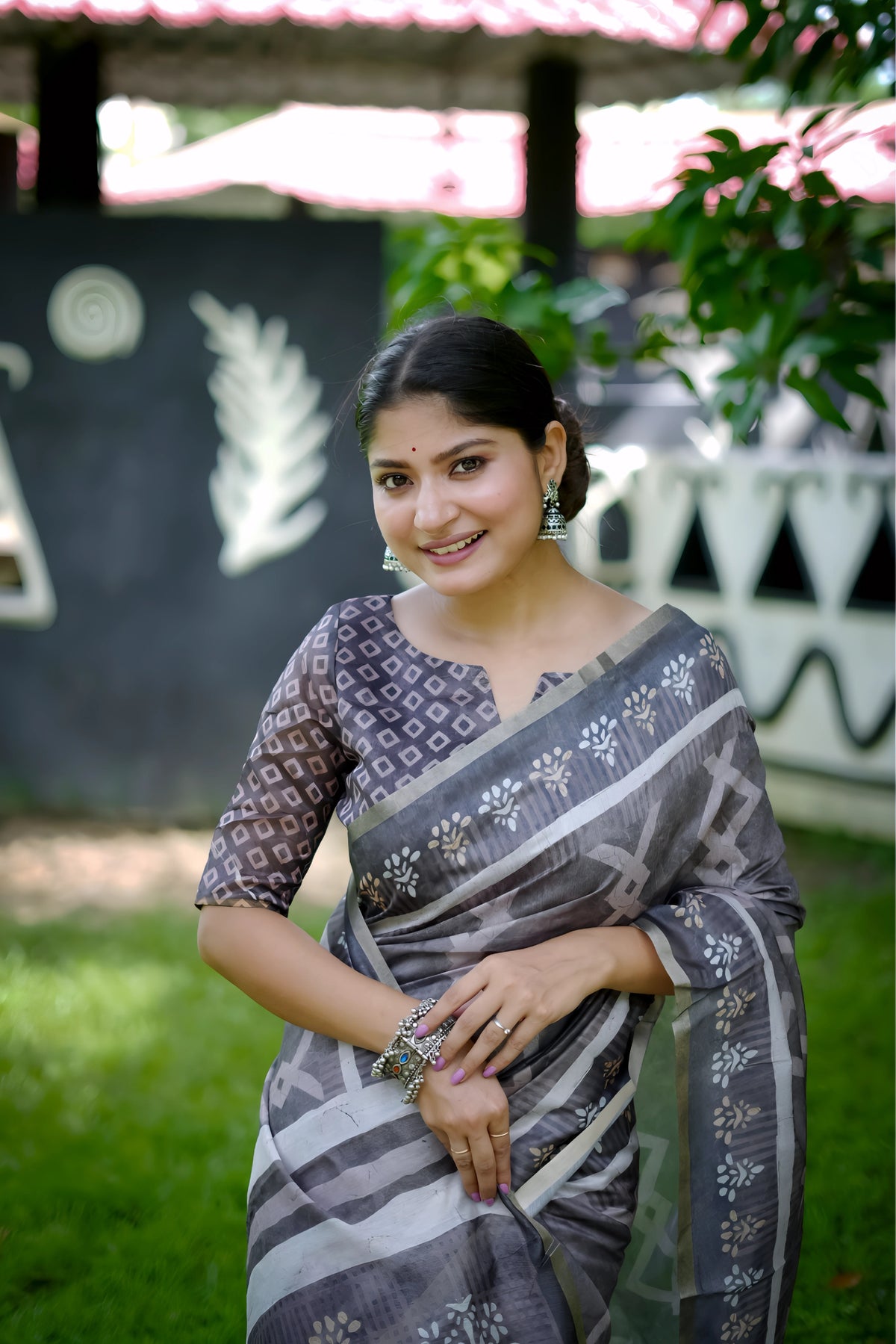 A woman models a striking Charcoal Grey Soft Cotton Saree featuring an all-over digital block print of geometric and abstract motifs in white. She wears a printed grey blouse and stacked silver bangles, posing outdoors against a stylized black and white wall.