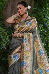 A woman models a sophisticated Slate Grey Soft Tussar Silk Saree. The saree features a wide pallu with a contrasting light yellow Kalamkari-style print, decorated with large floral motifs in dusty pink, blue, and green. She wears a matching short-sleeve printed blouse and accessorizes with heavy oxidized silver jewelry, posing outdoors.