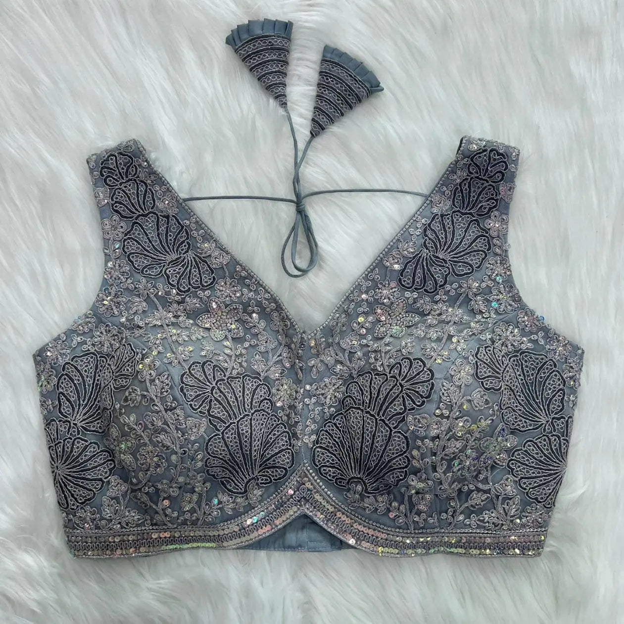 Elegant grey blouse featuring intricate sequin and thread embroidery with floral and leaf motifs, deep V-neck with structured cups, geometric border, and tasseled tie-back detail, displayed flat on a white fur background.