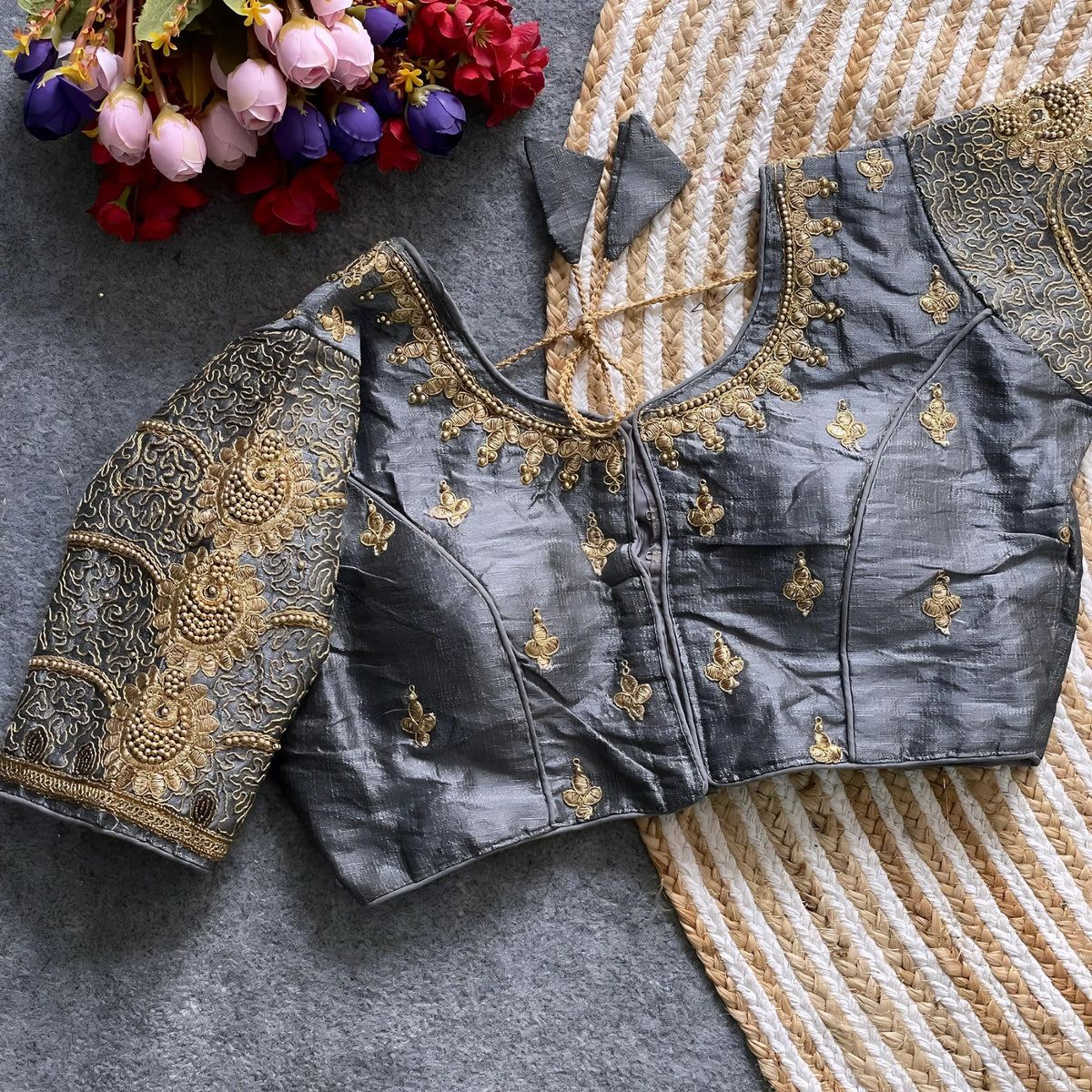 Traditional grey silk blouse featuring intricate gold embroidery and beadwork with floral and paisley motifs on the sleeves and neckline, displayed flat on a textured surface with a bunch of artificial flowers and a woven mat in the background.