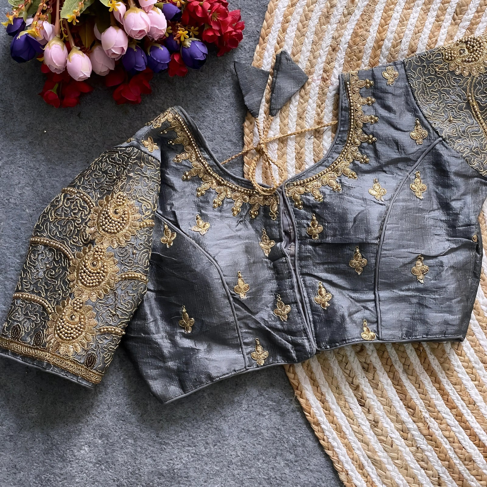 Traditional grey silk blouse featuring intricate gold embroidery and beadwork with floral and paisley motifs on the sleeves and neckline, displayed flat on a textured surface with a bunch of artificial flowers and a woven mat in the background.