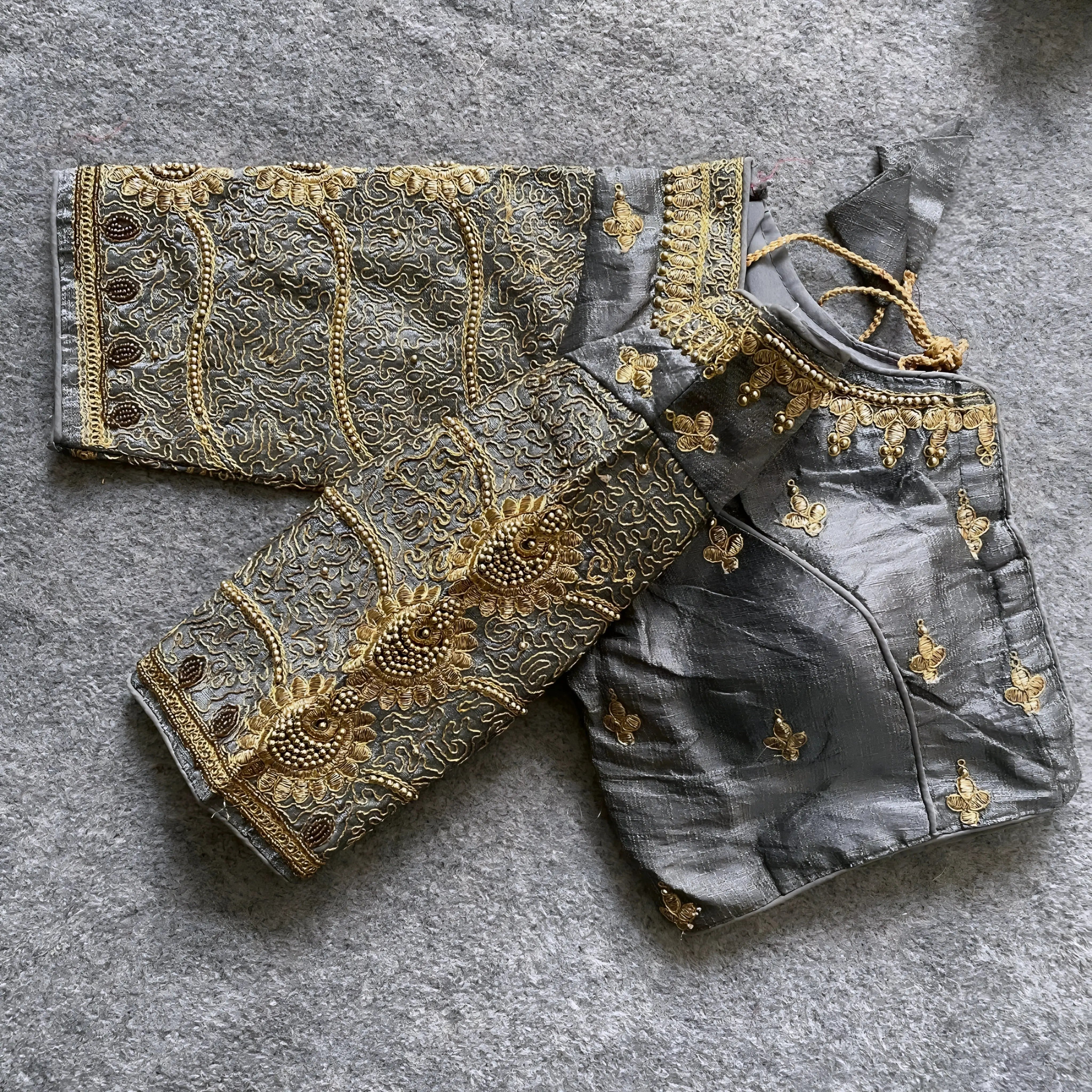 Traditional grey silk blouse featuring intricate gold embroidery and beadwork with floral and paisley motifs on the sleeves and neckline, displayed flat on a textured surface with a bunch of artificial flowers and a woven mat in the background.