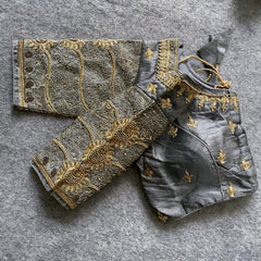 Traditional grey silk blouse featuring intricate gold embroidery and beadwork with floral and paisley motifs on the sleeves and neckline, displayed flat on a textured surface with a bunch of artificial flowers and a woven mat in the background.