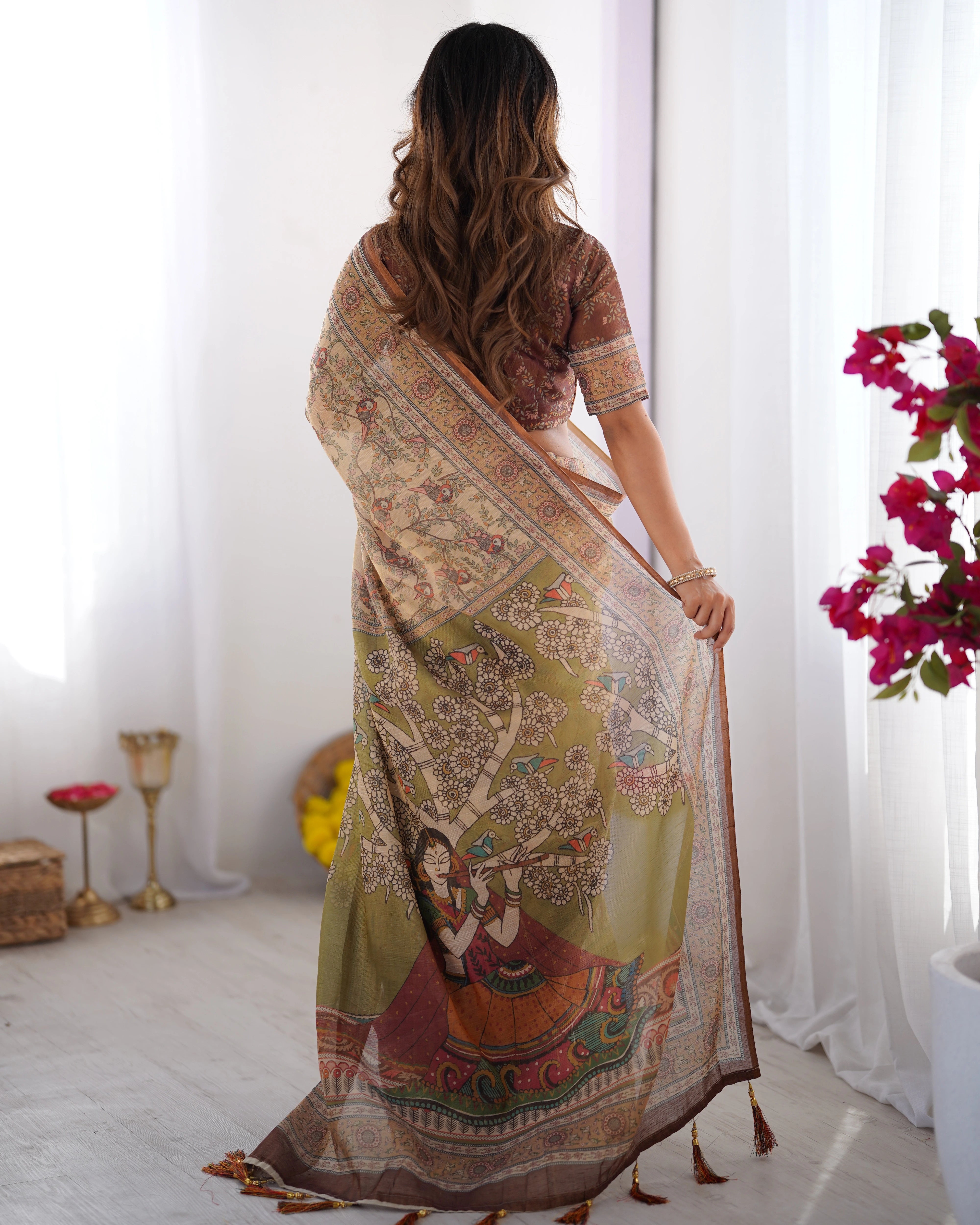 Woman wearing a patterned saree in a bright room with flowers and decor items.