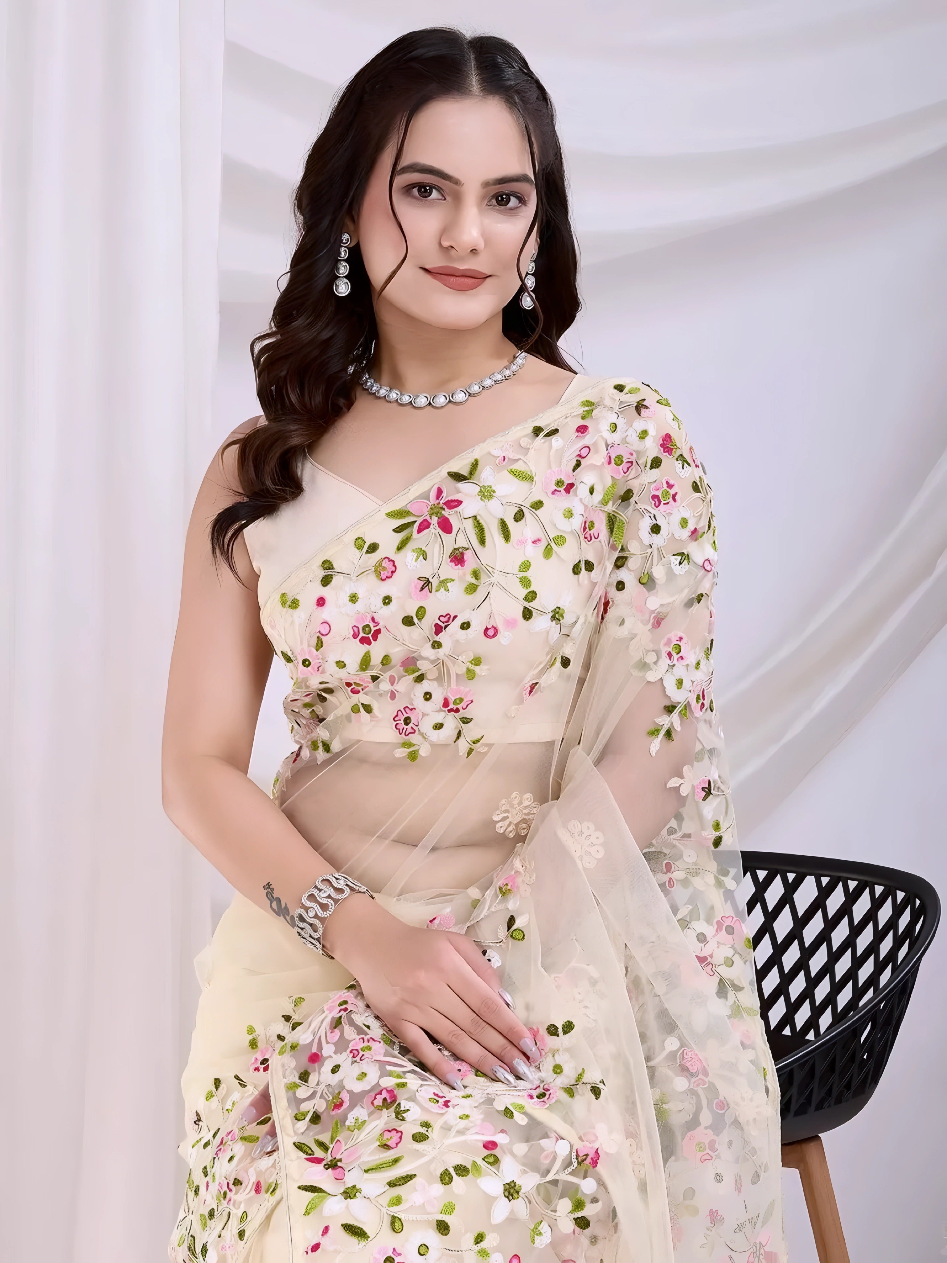 A woman models an ethereal Light Beige Soft Net Saree. The sheer fabric is densely covered in delicate all-over floral embroidery featuring white, pink, and green threadwork. She wears a matching sleeveless embroidered beige blouse and accessorizes with sophisticated silver/diamond jewelry, including a layered necklace and cuff bracelet.