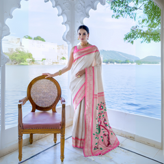 A woman models an elegant White or Cream Soft Silk Saree. The saree features a subtle tone-on-tone weave and a striking pink contrast border with thin gold Zari. The pallu is a focal point, featuring rich, artistic Paithani-style print or weaving of pink, red, and green bird-on-branch motifs. She wears a vibrant pink sleeveless blouse and gold Kundan jewelry, posing outdoors by a lake.