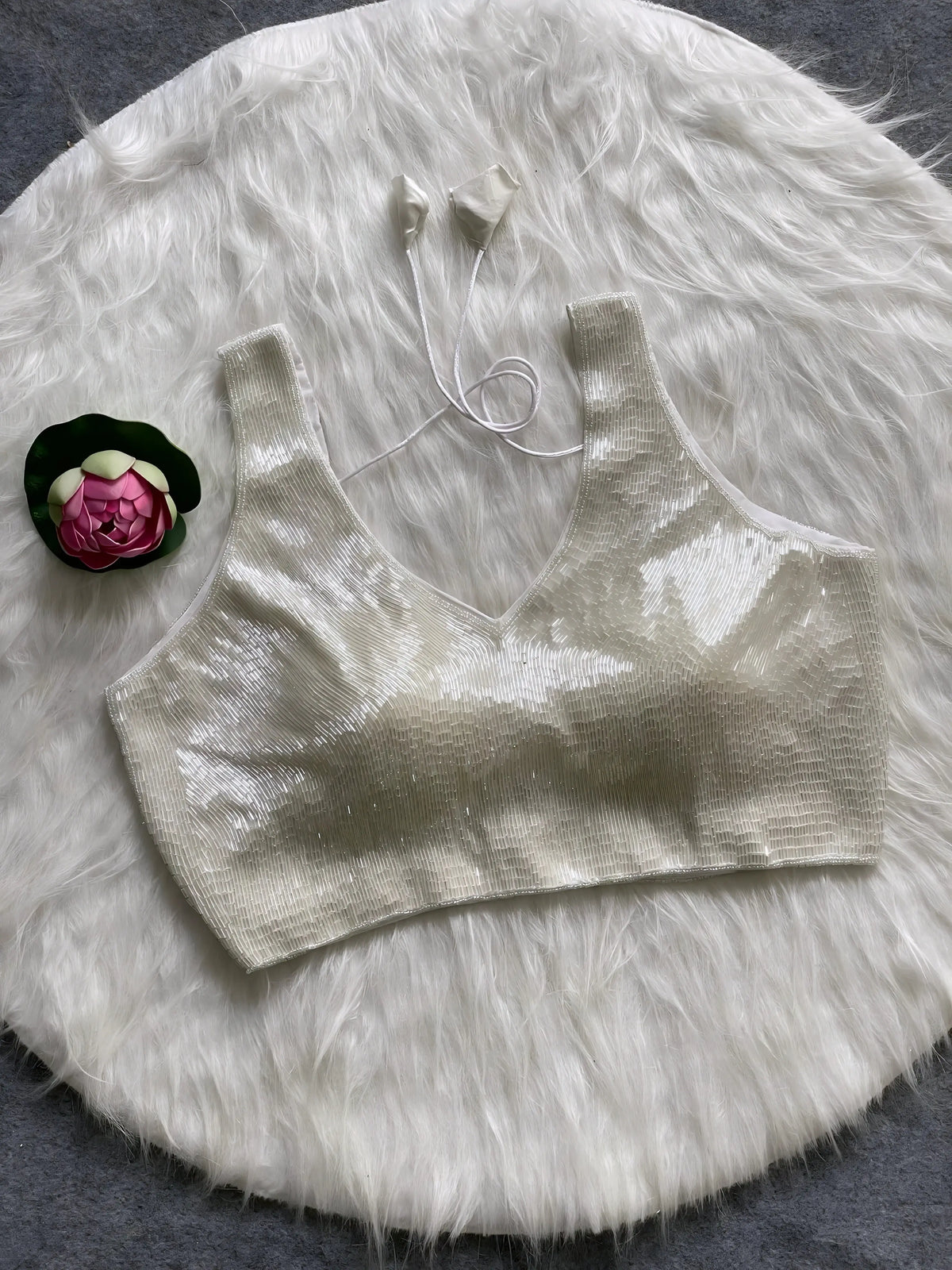 Ivory sequin sleeveless blouse with tie-back detail, displayed on a white fur background with a pink floral prop on the side